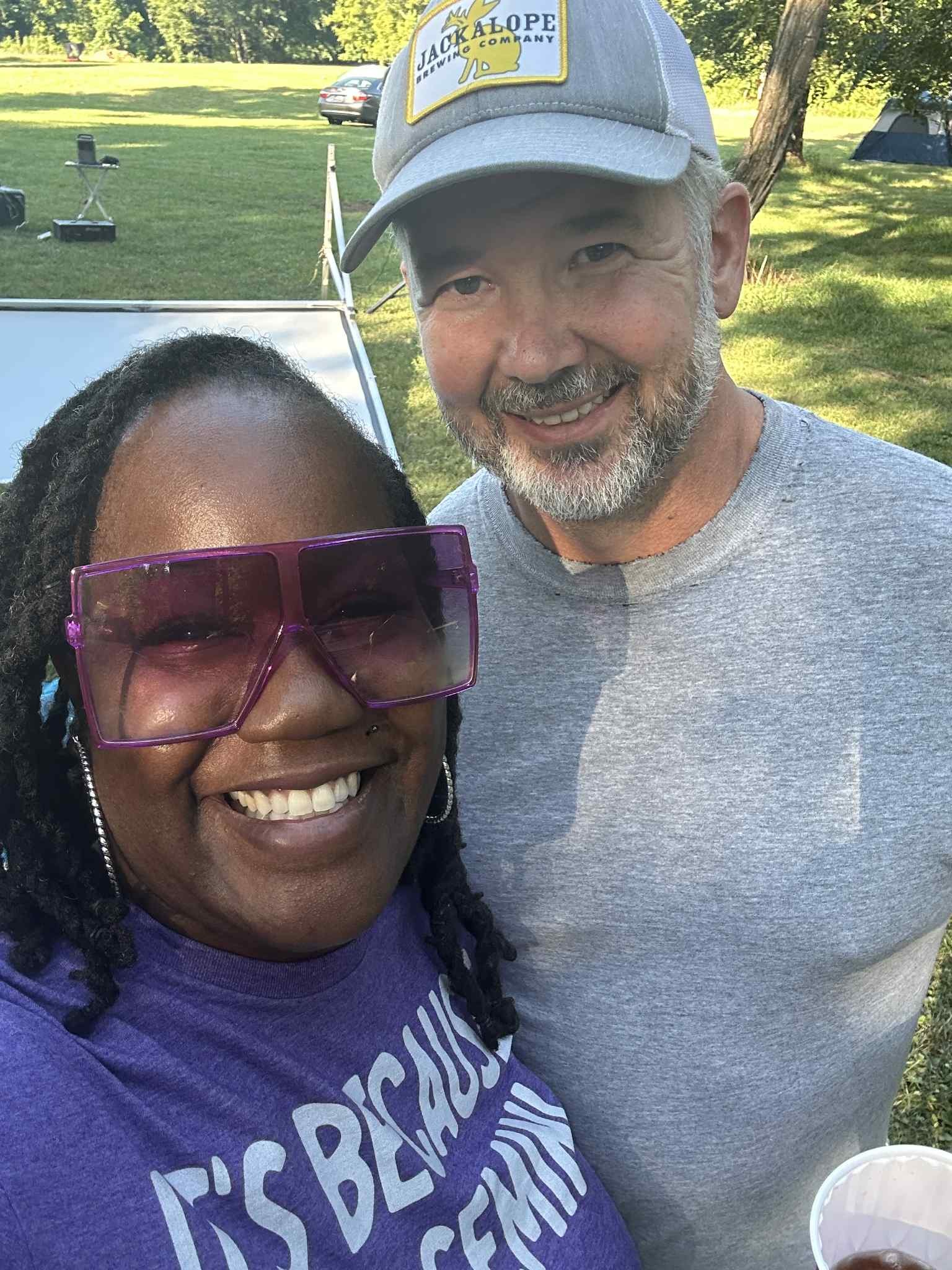 A couple smiling together in the wooded backyard on a sunny afternoon — Campout 2025