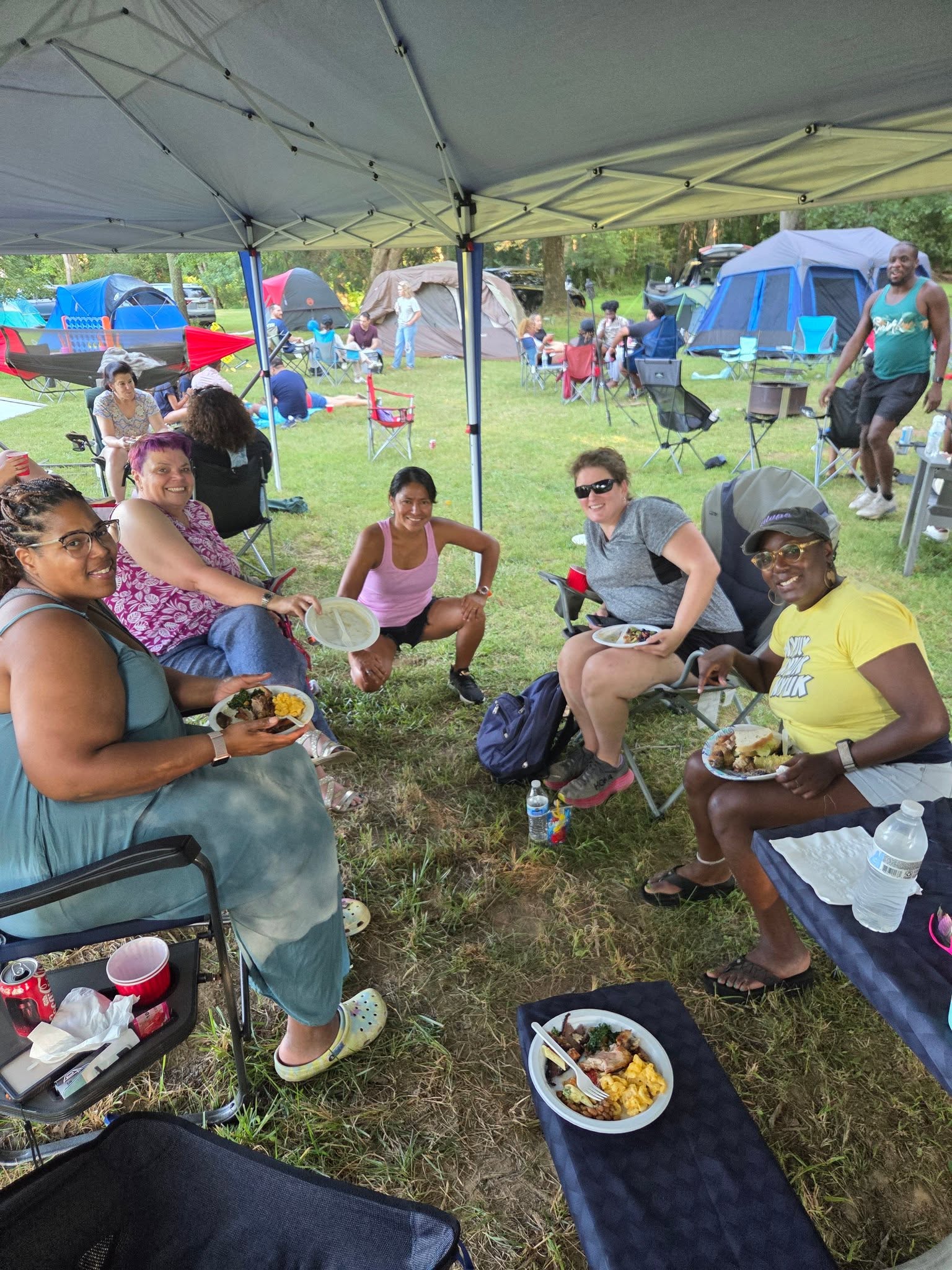 Guests eating and laughing under the canopy with tent city behind — Campout 2025