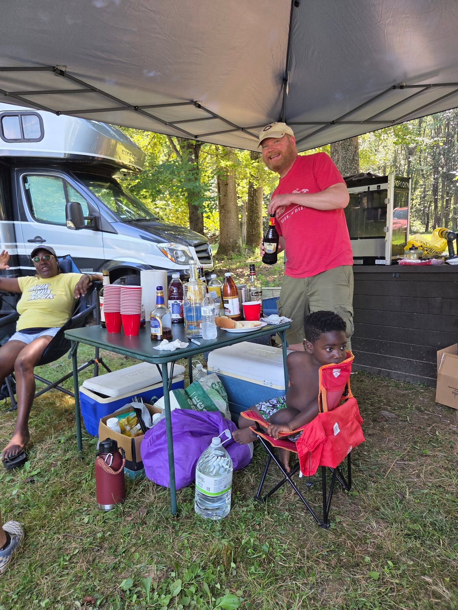 At the drink station under the canopy, child nearby and the RV behind — Campout 2025