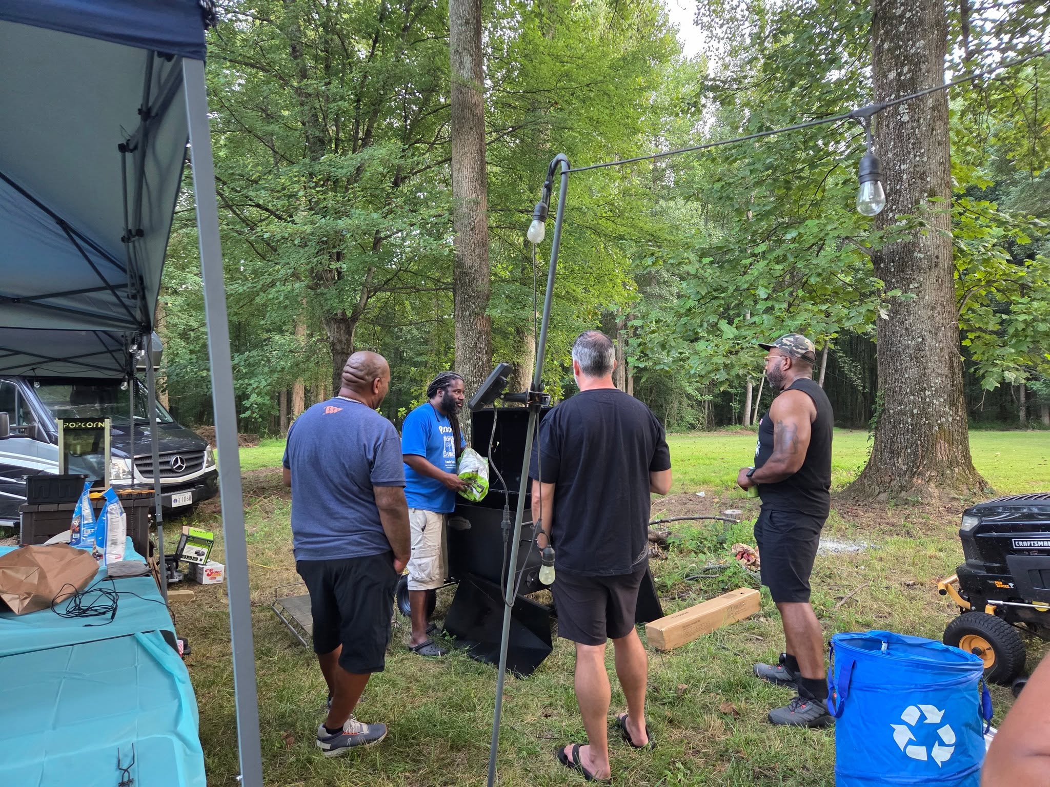 Guests gathered around the smoker in the wooded backyard during setup — Campout 2025