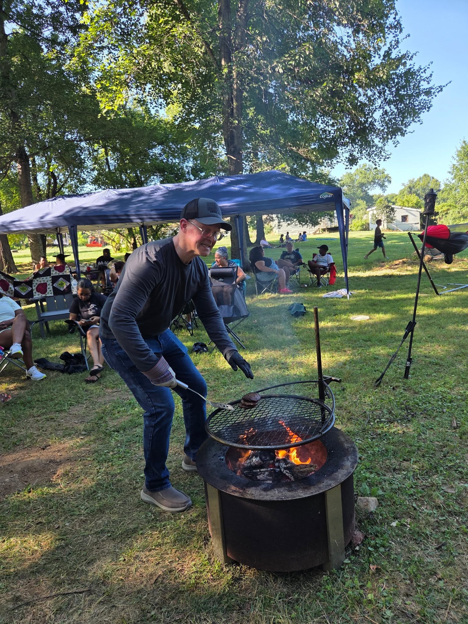 Tending a cast-iron pot over an open campfire with guests seated behind — Campout 2025